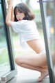 A woman in a white shirt and panties leaning against a window.