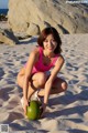 A woman in a pink bathing suit crouching on a beach with a coconut.