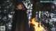 A woman in a black cloak standing in front of a fire.